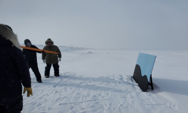 One person prepares to throw harpoon at a board painted with two black humps to symbolize a whale. Two other people watch.