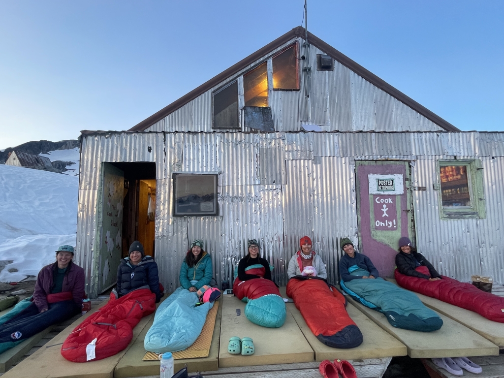 Seven people sit up in sleeping bags in front of a building
