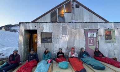 Seven people sit up in sleeping bags in front of a building