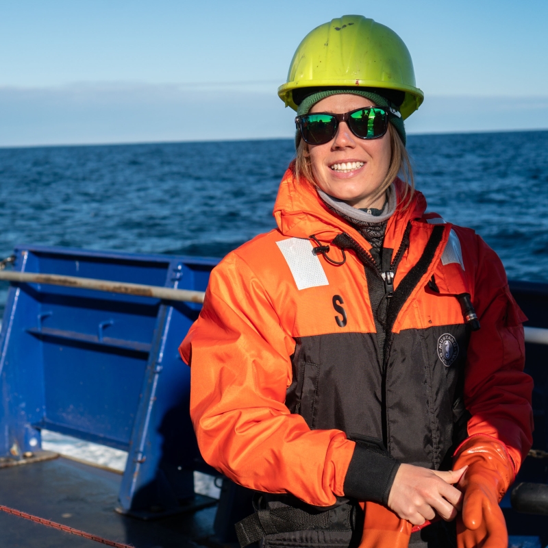 A woman smiling on a boat wearing a hardhat, sunglasses, a jacket, and gloves