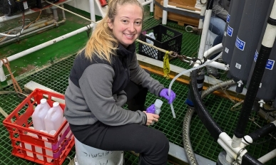 Kiley smiling at the camera while sitting on a bucket. She is holding a tube that is pulling water out of a larger machine. She holds a bottle for the water sample in her other hand.