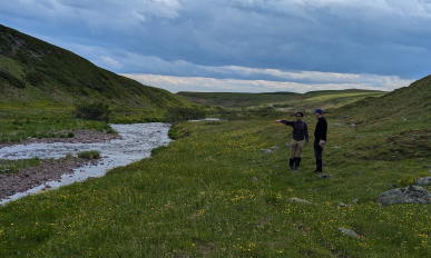 Two people stand in a field of grass. One of them points at a stream of water running nearby.