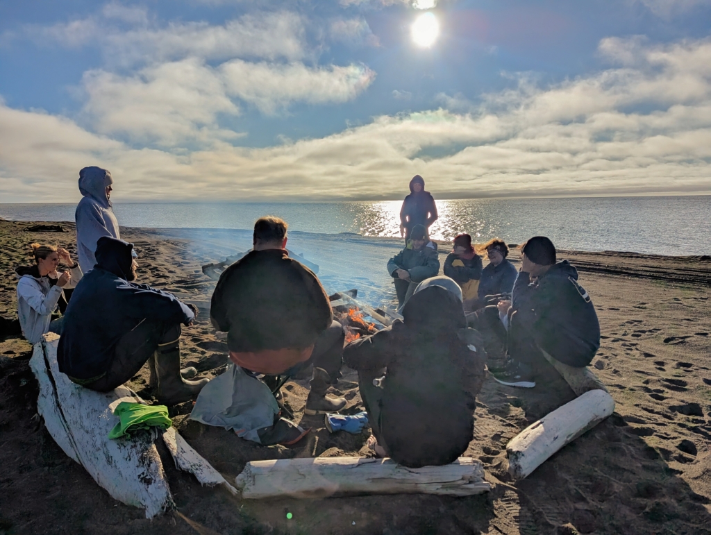 A group of people sits on the sand around a fire pit