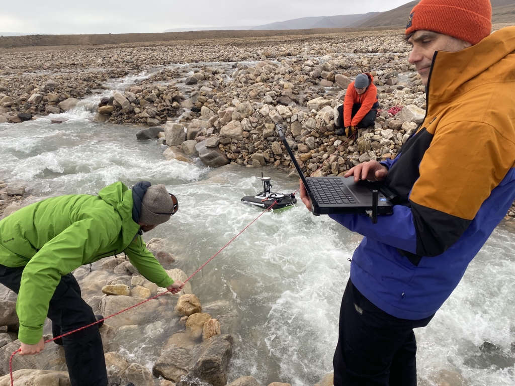 Three people stand on the edge of a stream. Two are holding rope attached to a machine in the stream and one is holding and looking at a laptop.