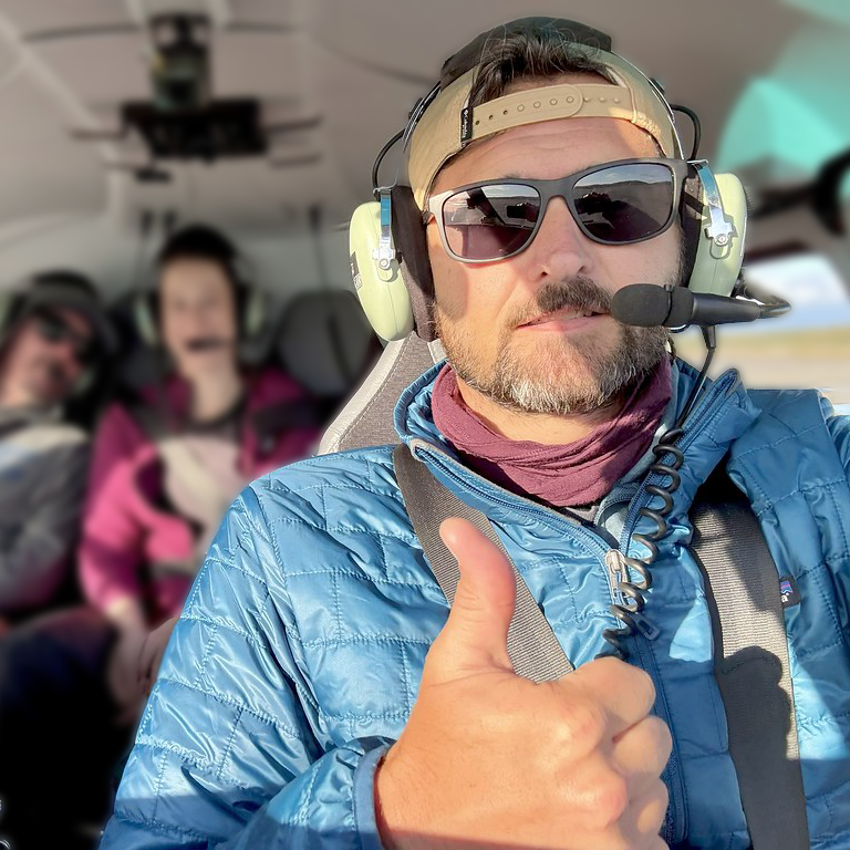 James sitting in a plane wearing a mic and headset. He is giving a thumbs up to the camera.