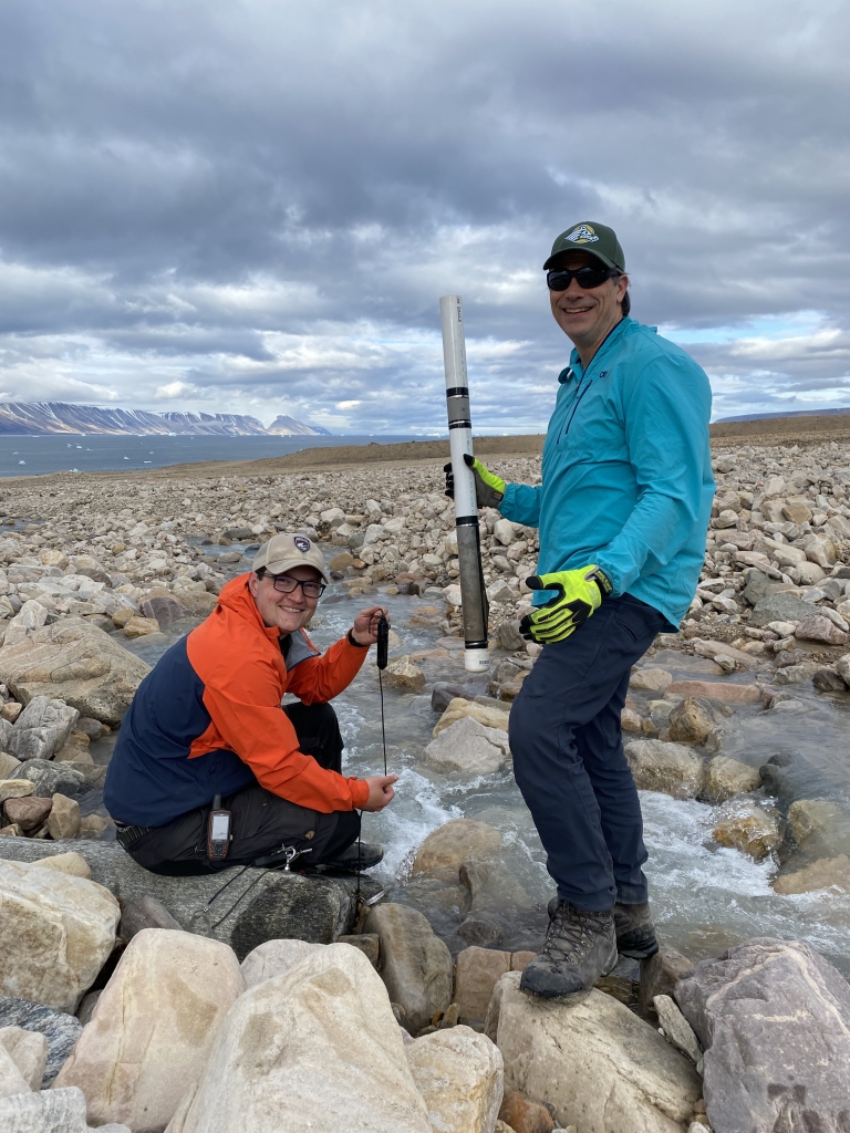 One person stands while another crouches on rocks near a stream. They are both smiling and holding equipment.