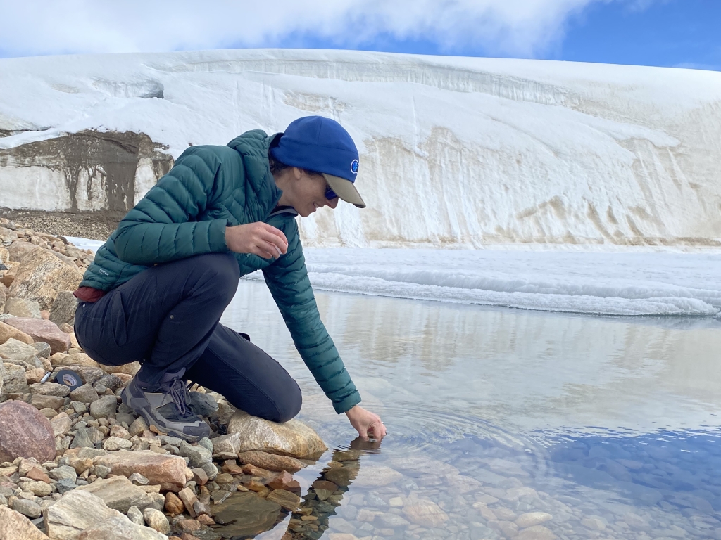 A woman reaches into the stream to collect a water sample