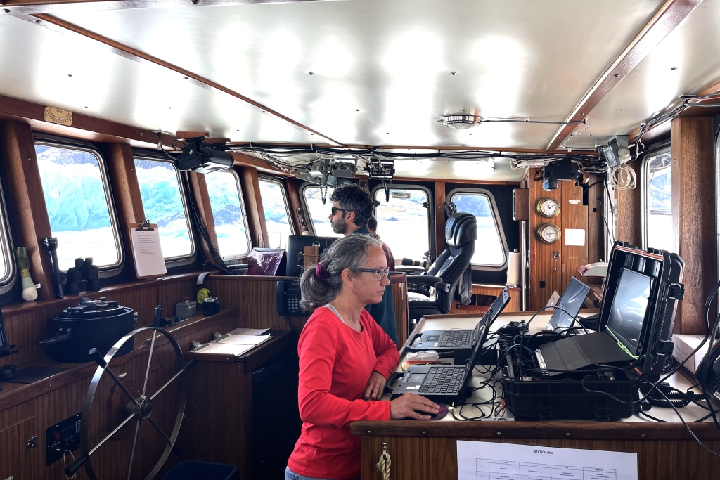 Two people standing inside a boat, one looks at a computer the other looks out the window