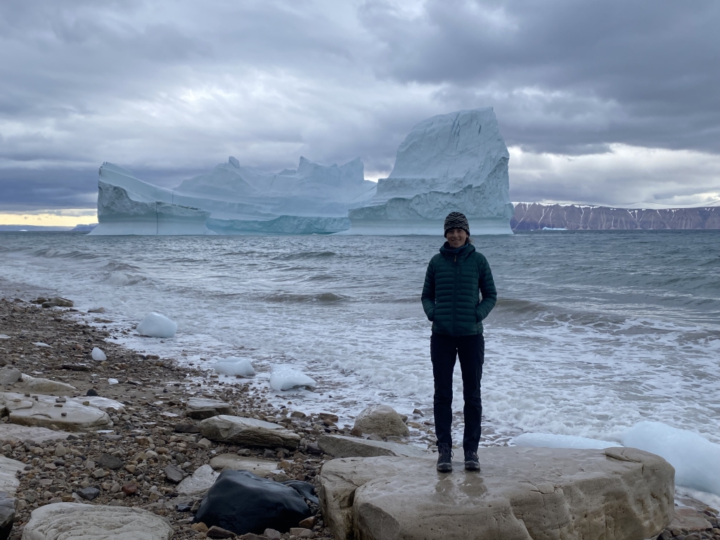 A woman stands on a rock at the shore of the sea. A large iceberg is behind her in the background.