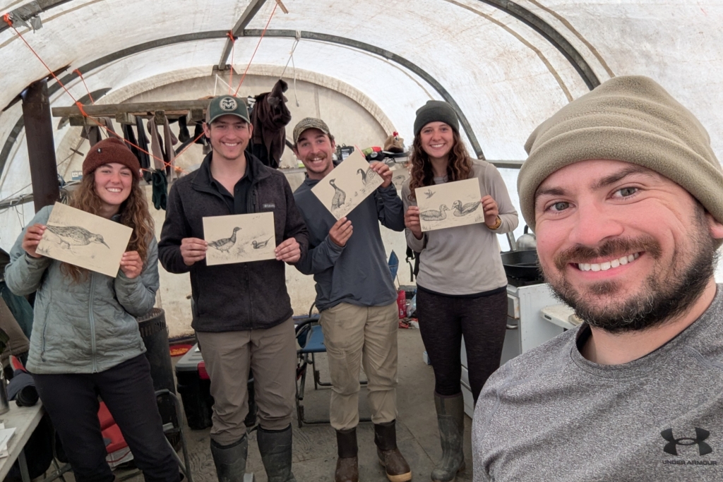 A man smiling at the camera, in the background are four people holding up drawings of birds and smiling