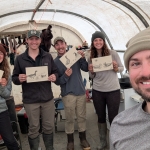 A man smiling at the camera, in the background are four people holding up drawings of birds and smiling