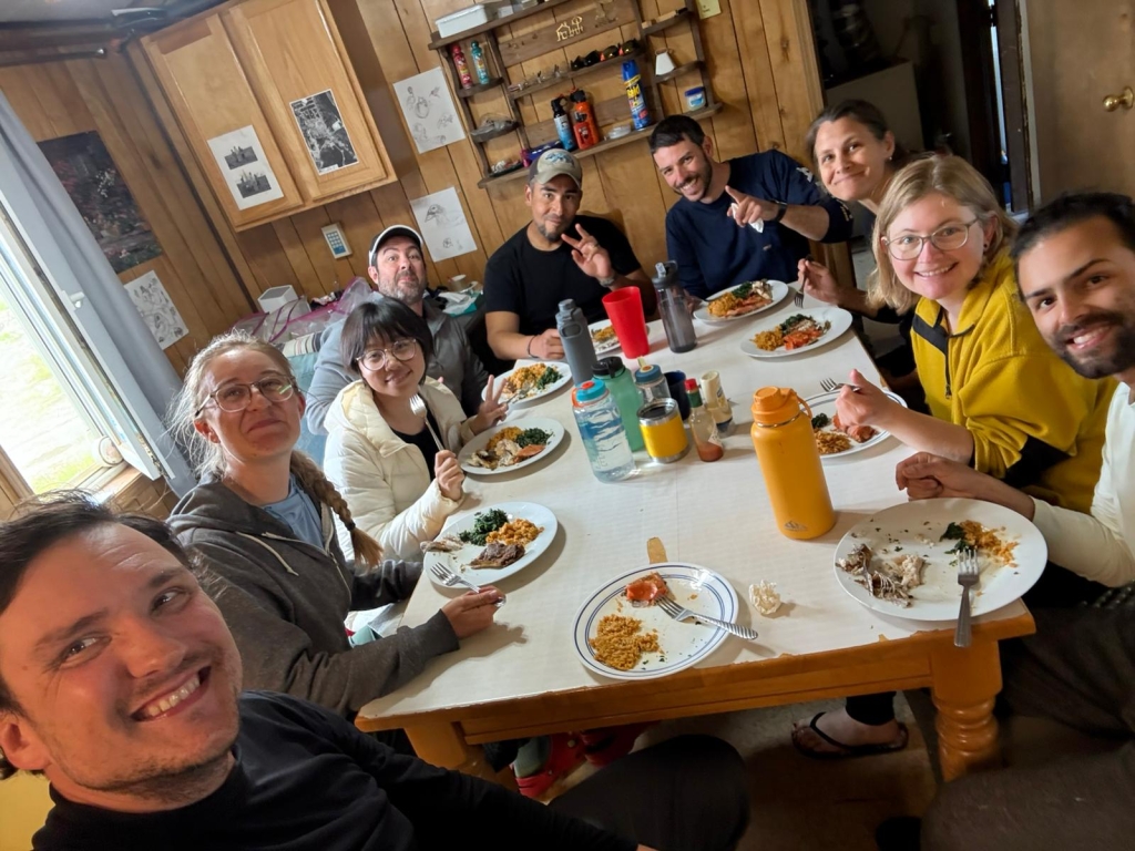 A group of people sit at a table with plates of food, they are facing the camera and smiling