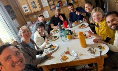 A group of people sit at a table with plates of food, they are facing the camera and smiling
