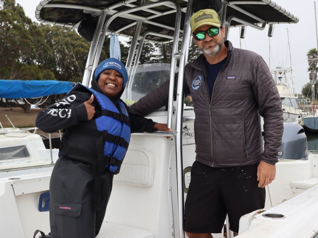 A woman stands on a boat smiling and giving a thumbs up while a man stands next to her smiling at the camera 