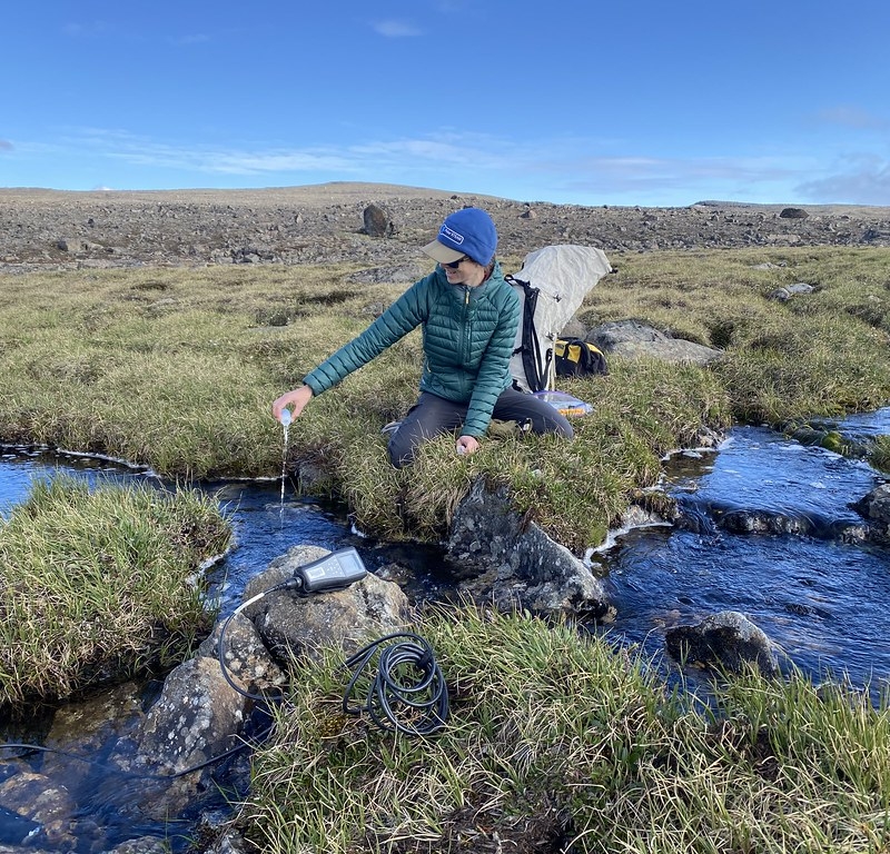 A woman holds a container with water pouring out of it back into a stream