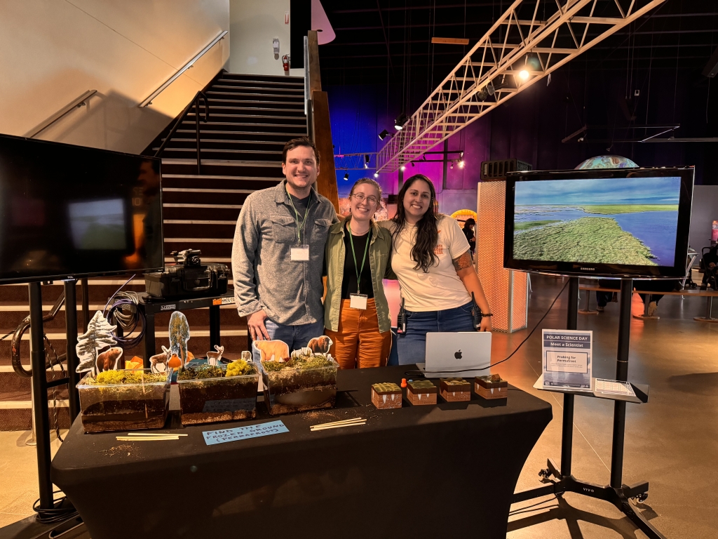 Three people smiling behind a table. A sign near them says Polar Science Day Meet a Scientist