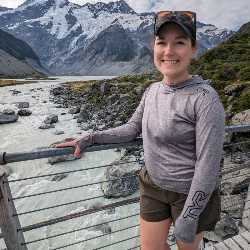 Kathleen stands on a bridge smiling there is a mountain in the background