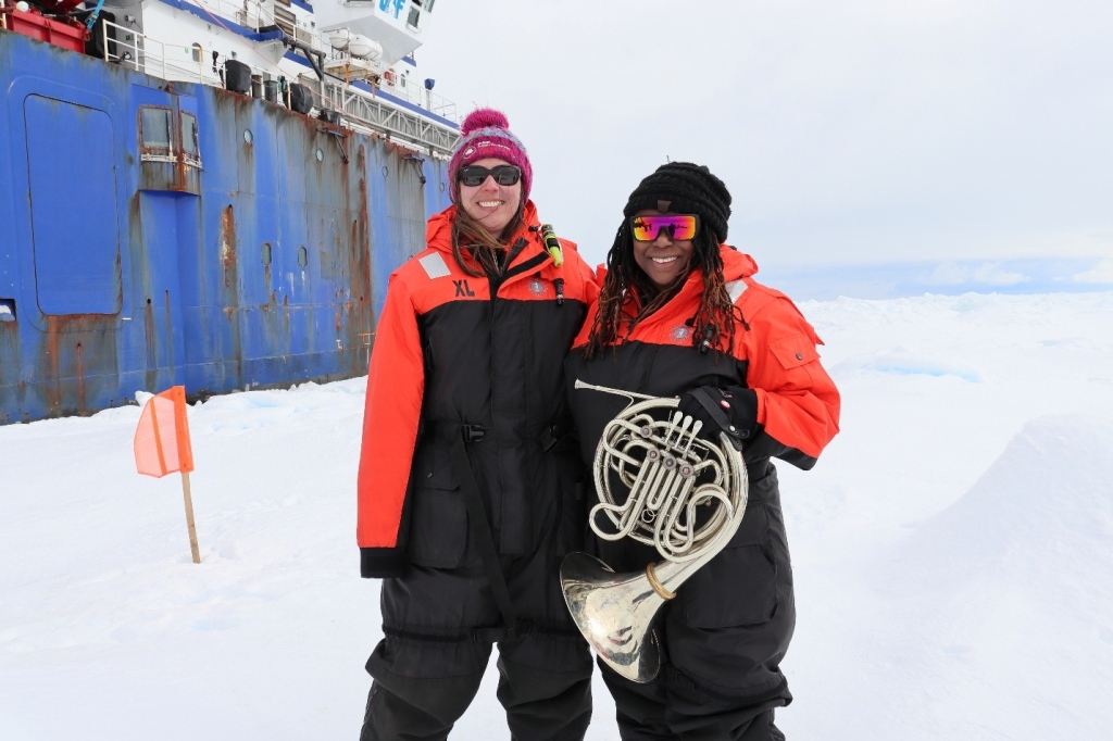 A woman researcher standing on ice shelf with woman artist holding a french horn. Blue hulled research ship in the background.