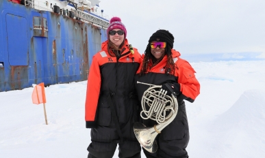 A woman researcher standing on ice shelf with woman artist holding a french horn. Blue hulled research ship in the background.