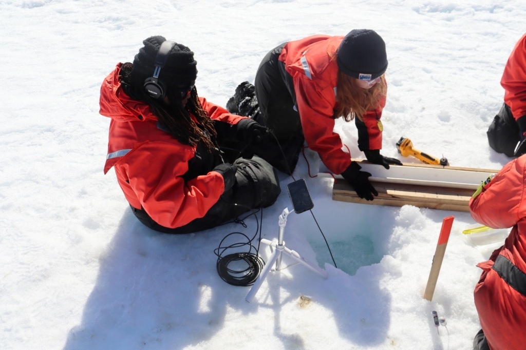 Woman artist sitting on the sea ice with headphones using a hydrophone. Also pictured woman scientist collecting sea ice data.
