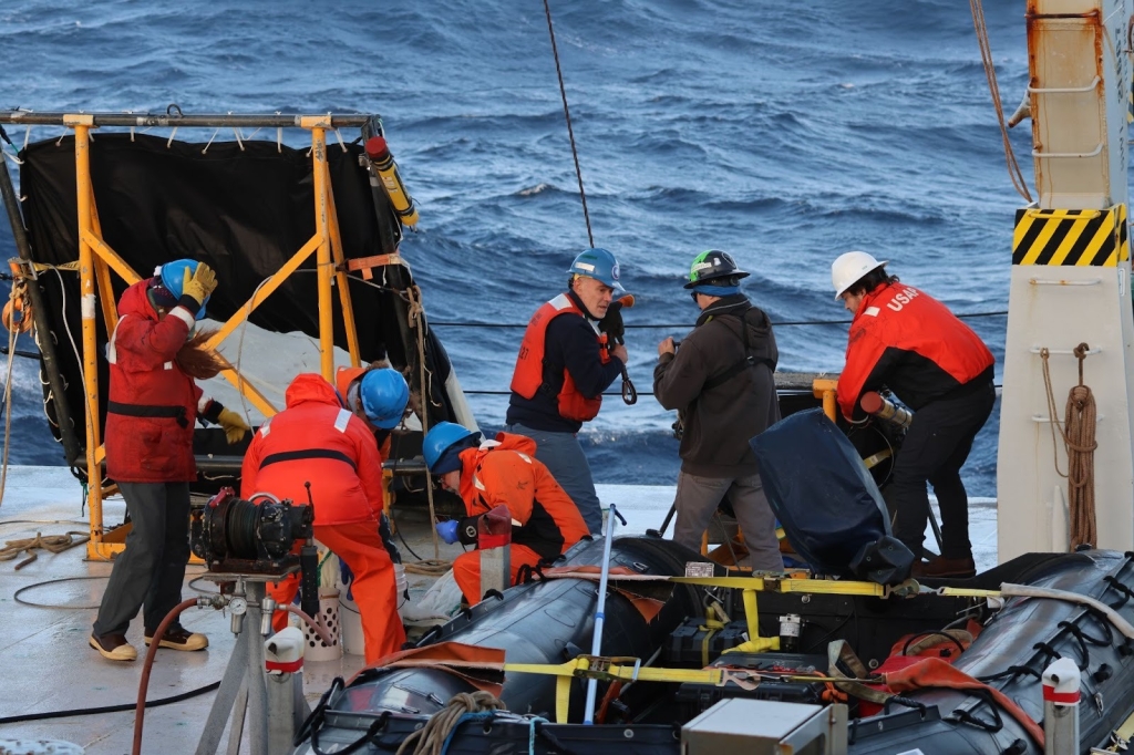 Six people work with nets and research equipment at the edge of a ship