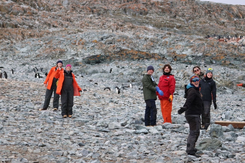7 people smile and pose in rocky terrain with many penguins in the background