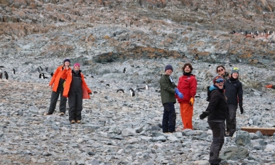 7 people smile and pose in rocky terrain with many penguins in the background