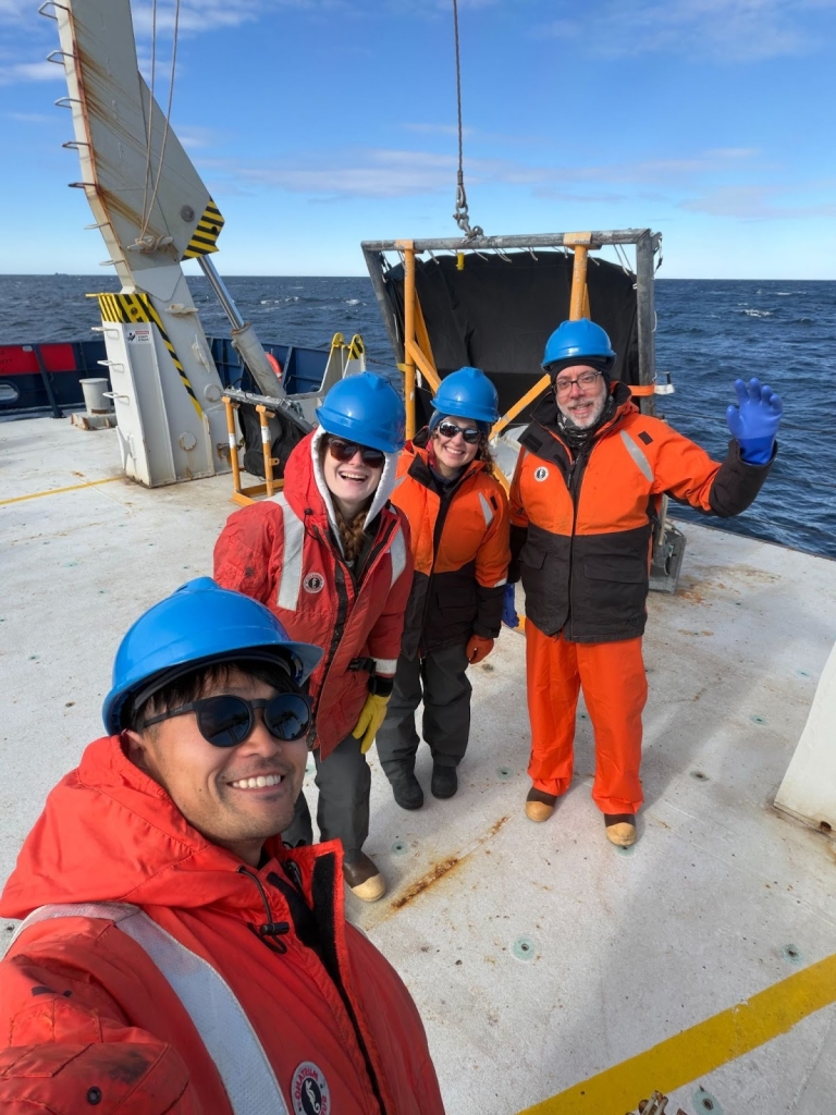Four people smiling and wearing cold weather gear and helmets at the edge of a ship