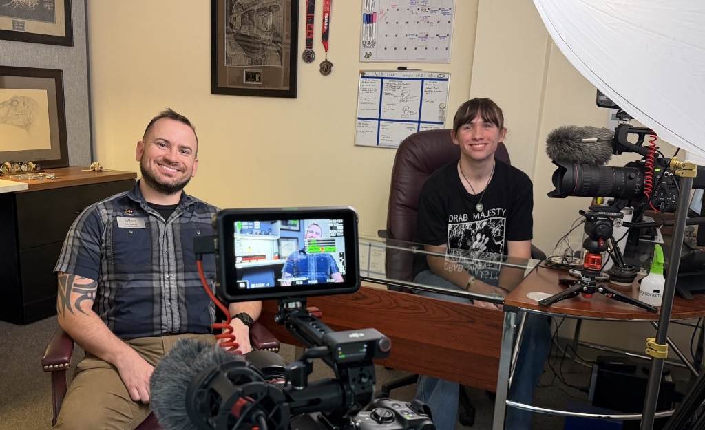 Two people smiling in an office with cameras pointed towards them
