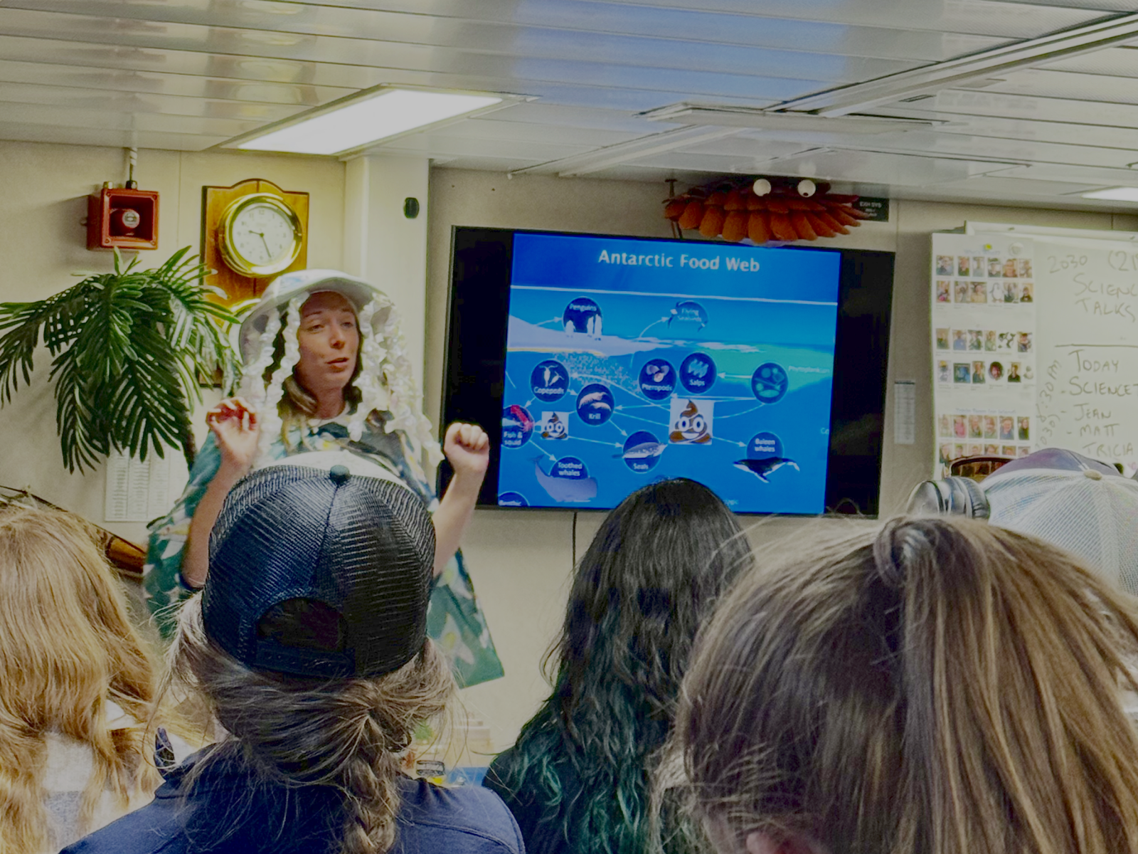 A person stands in front of a four seated people wearing a hat that looks like a jellyfish. Behind them on a TV screen is a slide that says "Antarctic Food Web".