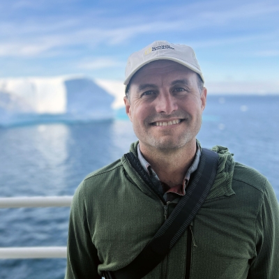 Andrew standing on a ship with iceberg and blue water in the background