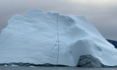 Large iceberg floating in calm water with mountainous land in the background under a cloudy sky
