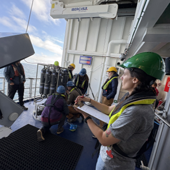 A person holding a clipboard while others work in the background all of whom are wearing safety vests and helmets.