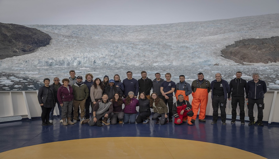 A group of people posing on a ship deck with a large glacier and rocky terrain in the background. The group wears outdoor clothing, including jackets and waterproof gear.