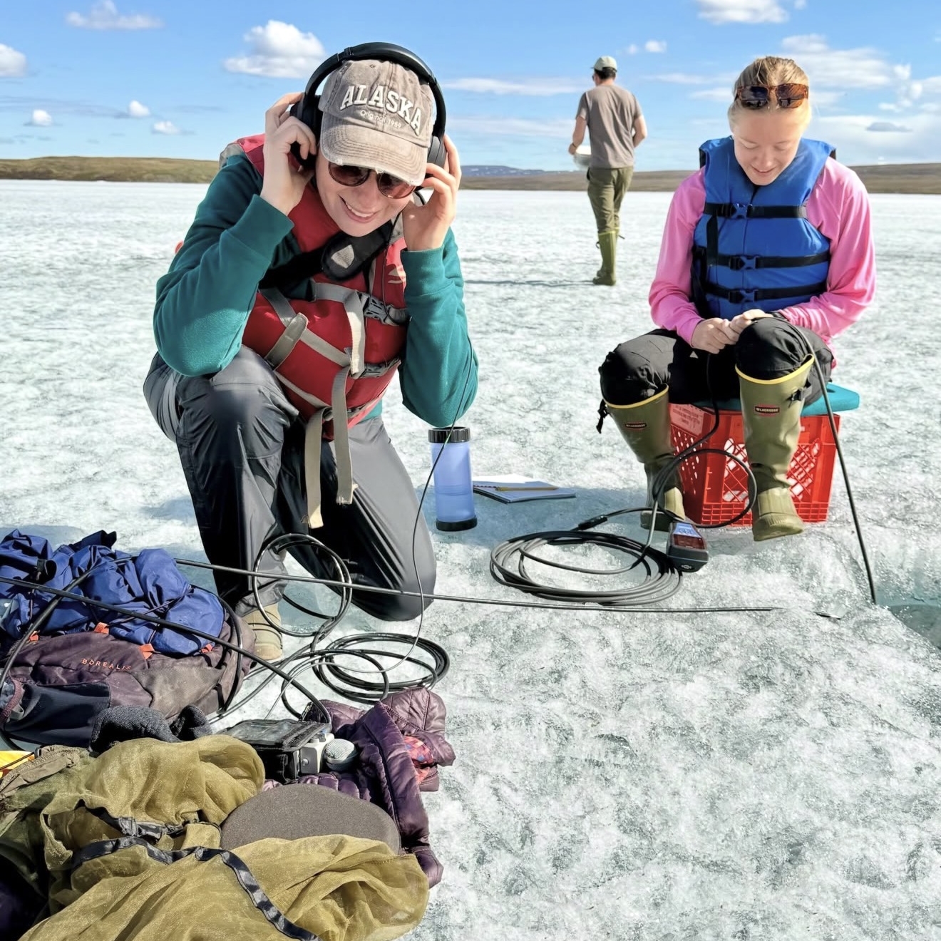 Two people sit on the ice with wires that go into a hole in the ice