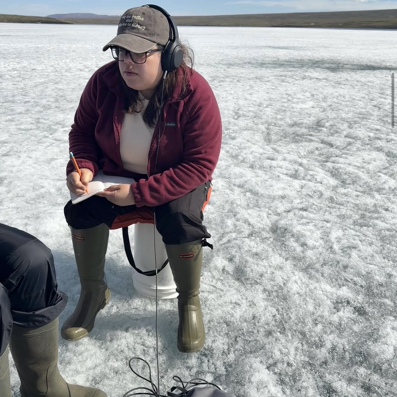 Karla takes notes in a notebook while sitting on a bucket on the ice and wearing headphones