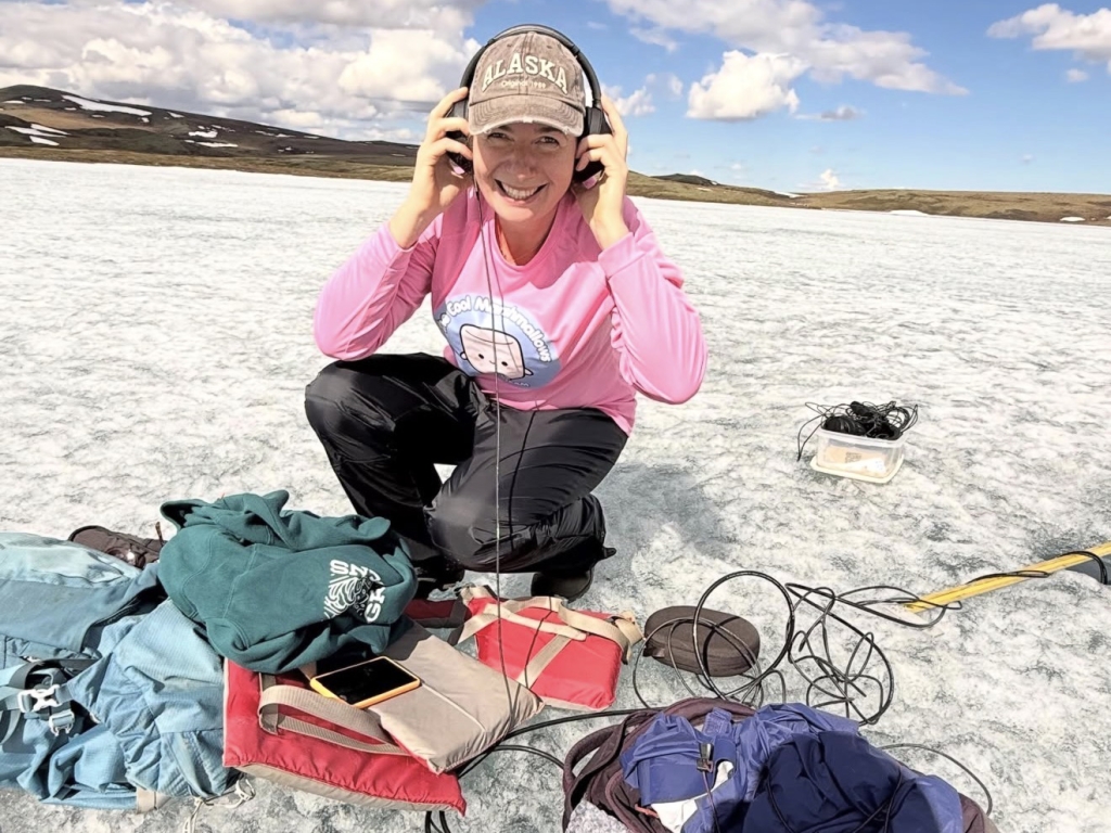 A person kneeling on ice and smiling at the camera while listening to something through headphones