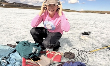 A person kneeling on ice and smiling at the camera while listening to something through headphones