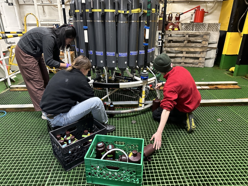 Three people collecting water out of a large collection of tanks into smaller containers via rubber hoses